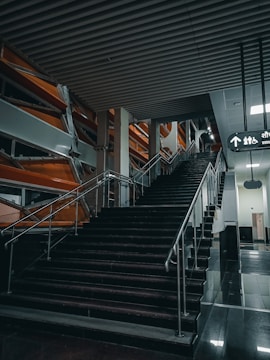 A modern stairway with black steps and metal railings in an indoor setting. The stairway leads to an upper level with a sign above indicating directions and symbols for an elevator and restroom. The surrounding area has an industrial design with orange and silver structural elements and a ceiling with horizontal beams.