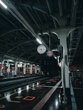 An empty train station platform with a clock hanging from the ceiling, showing the time as nearly 11:10. The platform is well-lit with fluorescent lights, and the area is clean with reflective floors. Signage with the number two is visible, indicating platform number. The overall atmosphere is quiet and still, suggesting nighttime.