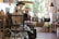 A quaint boutique interior featuring a variety of items including jewelry, mannequins dressed in elegant clothing, and furniture with decorative pillows. The store is well-lit with a mix of natural light from large windows and ambient lighting fixtures. Displays are neatly organized with attention to detail.