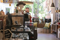 A quaint boutique interior featuring a variety of items including jewelry, mannequins dressed in elegant clothing, and furniture with decorative pillows. The store is well-lit with a mix of natural light from large windows and ambient lighting fixtures. Displays are neatly organized with attention to detail.