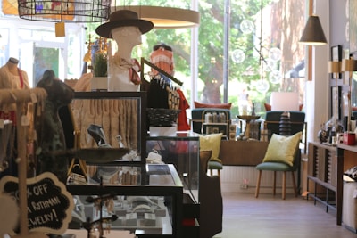 A quaint boutique interior featuring a variety of items including jewelry, mannequins dressed in elegant clothing, and furniture with decorative pillows. The store is well-lit with a mix of natural light from large windows and ambient lighting fixtures. Displays are neatly organized with attention to detail.