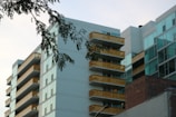 Modern apartment buildings with glass facades and yellow balconies are seen from below. A branch with green leaves hangs in the foreground, set against a partly cloudy sky.