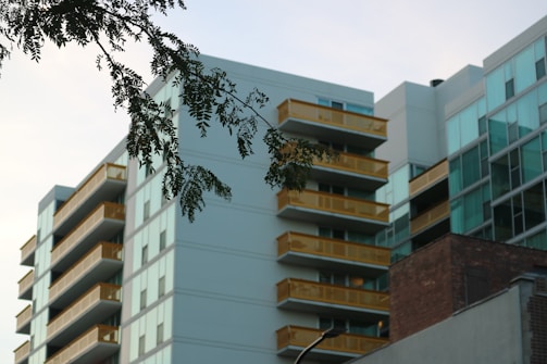 Modern apartment buildings with glass facades and yellow balconies are seen from below. A branch with green leaves hangs in the foreground, set against a partly cloudy sky.