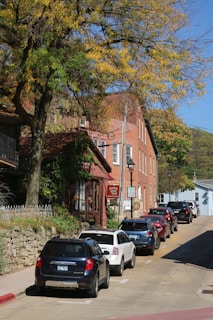 A quaint street scene featuring parked cars lined along a sidewalk under a large tree with autumn foliage. The buildings have a rustic charm with brick facades, and a storefront sign reading 'Dining House Tours' is visible. The atmosphere is serene, suggesting a small, historic town.