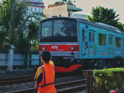 A person wearing an orange safety vest stands near a railway track, facing an approaching commuter train. Tall palm trees and urban buildings are visible in the background, suggesting an urban setting.