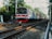 A commuter train travels along a railway track surrounded by lush greenery and urban structures. The train is seen moving towards the viewer, with multiple carriages painted in different colors. Overhead power lines are visible against the sky, and there is some litter and debris near the tracks.