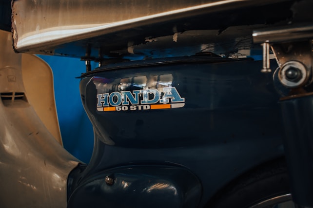 A close-up view of a vintage scooter's engine cover, displaying a Honda 50 STD emblem with metallic and orange accents under a metal structure.