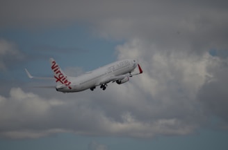 A commercial airplane with red and white livery is flying through a cloudy sky. The branding on the aircraft reads 'Virgin Australia.' The clouds are varied in density, some appearing rather dark, while others are lighter and thinner.