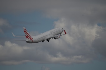 A commercial airplane with red and white livery is flying through a cloudy sky. The branding on the aircraft reads 'Virgin Australia.' The clouds are varied in density, some appearing rather dark, while others are lighter and thinner.