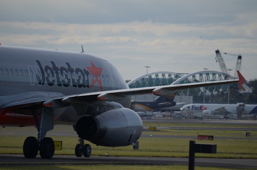 A commercial airplane with the logo on its fuselage is taxiing on a runway. In the background, other planes are visible, along with industrial structures like cranes and a bridge. The sky is overcast, with clouds scattered across.