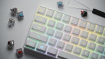 A mechanical keyboard with pastel multicolor backlighting is displayed diagonally across the image. Around the keyboard, several switch components and a keycap puller are scattered, indicating keyboard customization.