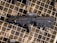 A black tactical firearm is displayed against a camouflaged patterned background. The gun features a compact design with a visible holographic sight mounted on top.