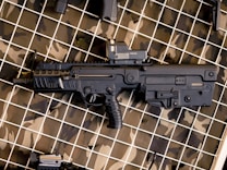 A black tactical firearm is displayed against a camouflaged patterned background. The gun features a compact design with a visible holographic sight mounted on top.