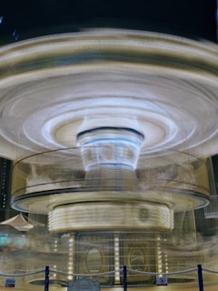 A blurred motion capture of a spinning carousel, with a glowing, luminous appearance. The structure appears to emit a warm light with circular patterns, and dense lines of light radiate outward, creating a dynamic, energetic effect.