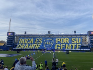 A large stadium with a prominent banner displaying the words 'BOCA ES GRANDE POR SU GENTE' in bold yellow and blue colors, featuring the emblem of CABJ in the center. Spectators and staff are visible around the field, with a clear blue sky in the background.