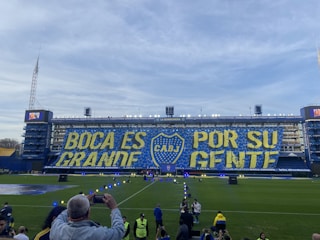 A large stadium with a prominent banner displaying the words 'BOCA ES GRANDE POR SU GENTE' in bold yellow and blue colors, featuring the emblem of CABJ in the center. Spectators and staff are visible around the field, with a clear blue sky in the background.