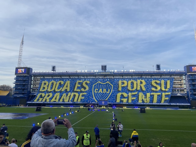 A large stadium with a prominent banner displaying the words 'BOCA ES GRANDE POR SU GENTE' in bold yellow and blue colors, featuring the emblem of CABJ in the center. Spectators and staff are visible around the field, with a clear blue sky in the background.