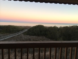 The image captures a serene beach scene with a wide, open view of the horizon where the sky meets the calm sea. The sky is painted in soft pastel hues of pink and blue, suggesting either sunrise or sunset. A wooden boardwalk leads through dense greenery and dunes, inviting a peaceful walk towards the shoreline.