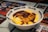 A bowl containing a creamy yellow omelet draped over rice, generously topped with a thick brown sauce. The bowl has 'SEEKER CLUB' written on its rim, and a shiny metal spoon rests inside. The background consists of menus with various images of food, hinting at a Japanese or Asian cuisine theme.