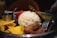 A metal tray contains a mound of white rice, a grilled sausage, grilled chicken, strips of bacon, tortilla chips, and a small metal cup. Some vegetables like lettuce and tomatoes are also present, accompanied by cutlery pieces.