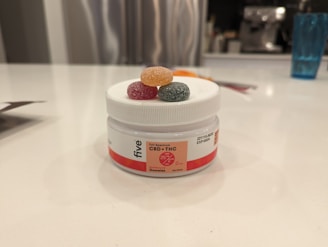 A white container labeled 'CBD + THC Gummies' with colorful gummy candies on top. The lid has three gummies in orange, red, and green. The setting is a modern kitchen with a blurred background featuring kitchen appliances and a blue glass.