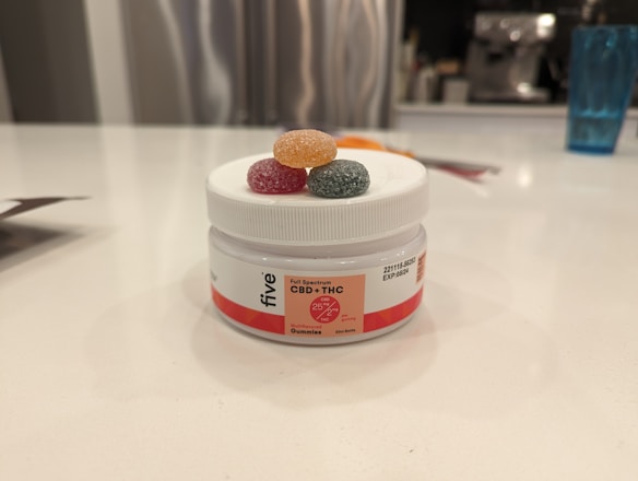 A white container labeled 'CBD + THC Gummies' with colorful gummy candies on top. The lid has three gummies in orange, red, and green. The setting is a modern kitchen with a blurred background featuring kitchen appliances and a blue glass.