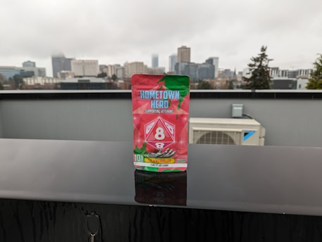 A package of ‘Hometown Hero’ stands prominently on a wet black surface, with a blurred cityscape featuring tall buildings and overcast skies in the background. The packaging design is vibrant with red and green colors, including images of watermelon slices. The text on the package highlights that it supports veterans and mentions ‘Delta 8’.
