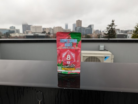 A package of ‘Hometown Hero’ stands prominently on a wet black surface, with a blurred cityscape featuring tall buildings and overcast skies in the background. The packaging design is vibrant with red and green colors, including images of watermelon slices. The text on the package highlights that it supports veterans and mentions ‘Delta 8’.