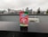A package of ‘Hometown Hero’ stands prominently on a wet black surface, with a blurred cityscape featuring tall buildings and overcast skies in the background. The packaging design is vibrant with red and green colors, including images of watermelon slices. The text on the package highlights that it supports veterans and mentions ‘Delta 8’.