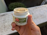 A hand holding a small white container labeled LivRelief Infused, featuring THC and CBD details. The label has a green background with a red THC symbol. The container is open, displaying a cream inside. The setting appears to be outdoors with a blurred background.
