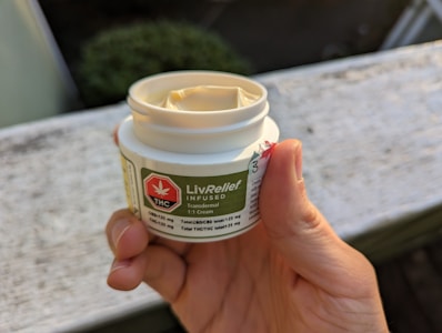 A hand holding a small white container labeled LivRelief Infused, featuring THC and CBD details. The label has a green background with a red THC symbol. The container is open, displaying a cream inside. The setting appears to be outdoors with a blurred background.