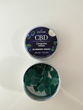 A tin of CBD gummies labeled 'Blueberry Dream' with an open lid revealing square-shaped green gummies inside. The label indicates it contains 25mg of CBD and is THC free. The tin has a dark blue label with images of blueberries and leaves.