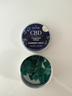A tin of CBD gummies labeled 'Blueberry Dream' with an open lid revealing square-shaped green gummies inside. The label indicates it contains 25mg of CBD and is THC free. The tin has a dark blue label with images of blueberries and leaves.