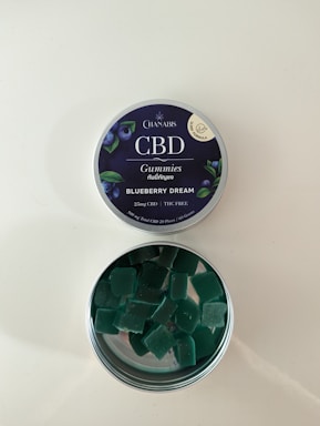 A tin of CBD gummies labeled 'Blueberry Dream' with an open lid revealing square-shaped green gummies inside. The label indicates it contains 25mg of CBD and is THC free. The tin has a dark blue label with images of blueberries and leaves.