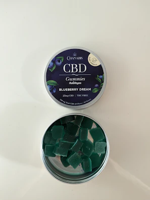 A tin of CBD gummies labeled 'Blueberry Dream' with an open lid revealing square-shaped green gummies inside. The label indicates it contains 25mg of CBD and is THC free. The tin has a dark blue label with images of blueberries and leaves.