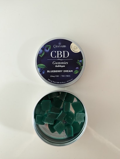 A tin of CBD gummies labeled 'Blueberry Dream' with an open lid revealing square-shaped green gummies inside. The label indicates it contains 25mg of CBD and is THC free. The tin has a dark blue label with images of blueberries and leaves.