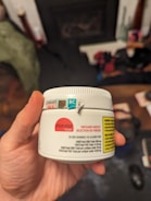 A hand holds a white container labeled with details about CBD gummies, including the brand 'Monjour Bare' and product information. The container features a label indicating cannabis content and a warning. In the background, there are blurred objects, possibly furniture or household items.