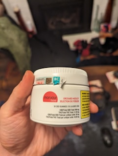 A hand holds a white container labeled with details about CBD gummies, including the brand 'Monjour Bare' and product information. The container features a label indicating cannabis content and a warning. In the background, there are blurred objects, possibly furniture or household items.
