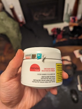 A hand holds a white container labeled with details about CBD gummies, including the brand 'Monjour Bare' and product information. The container features a label indicating cannabis content and a warning. In the background, there are blurred objects, possibly furniture or household items.