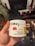A hand holds a white container labeled with details about CBD gummies, including the brand 'Monjour Bare' and product information. The container features a label indicating cannabis content and a warning. In the background, there are blurred objects, possibly furniture or household items.