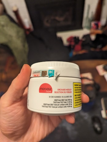 A hand holds a white container labeled with details about CBD gummies, including the brand 'Monjour Bare' and product information. The container features a label indicating cannabis content and a warning. In the background, there are blurred objects, possibly furniture or household items.