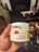 A hand holds a white container labeled with details about CBD gummies, including the brand 'Monjour Bare' and product information. The container features a label indicating cannabis content and a warning. In the background, there are blurred objects, possibly furniture or household items.
