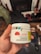 A hand holds a white container labeled with details about CBD gummies, including the brand 'Monjour Bare' and product information. The container features a label indicating cannabis content and a warning. In the background, there are blurred objects, possibly furniture or household items.