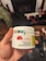 A hand holds a white container labeled with details about CBD gummies, including the brand 'Monjour Bare' and product information. The container features a label indicating cannabis content and a warning. In the background, there are blurred objects, possibly furniture or household items.