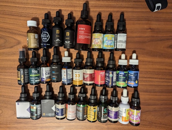 A collection of various tincture bottles arranged on a wooden surface. The bottles are primarily made of dark glass and feature dropper lids. Labels display different brands and product names related to CBD oil and tinctures. Some bottles have colorful labels with distinct branding, including text and imagery indicating different formulations.