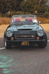 A classic Datsun convertible car is parked on a textured asphalt road. The car is in excellent condition, featuring a shiny dark green exterior with red interior seats. A customized California license plate reads 'SNICE 1'. The background features dry grass and tall trees, suggesting a rural setting.