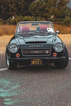 A classic Datsun convertible car is parked on a textured asphalt road. The car is in excellent condition, featuring a shiny dark green exterior with red interior seats. A customized California license plate reads 'SNICE 1'. The background features dry grass and tall trees, suggesting a rural setting.