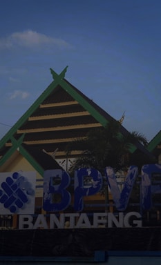 A large sign in front of a building with a triangular roof, featuring prominent blue and white lettering that reads 'BPVF Bantaeng.' The building in the background is topped with green trim and a palm tree is partially visible on the side.