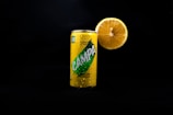 A can of lemon-flavored soda stands in a dark background with a large half of a lemon placed directly behind it. The can is covered in condensation droplets, indicating a cold temperature.