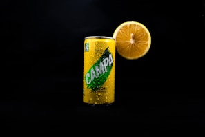 A can of lemon-flavored soda stands in a dark background with a large half of a lemon placed directly behind it. The can is covered in condensation droplets, indicating a cold temperature.
